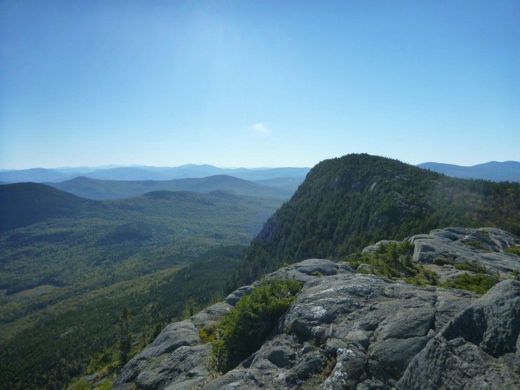 A view shows a distant mountain range and a nearby rocky mountain covered in green trees.