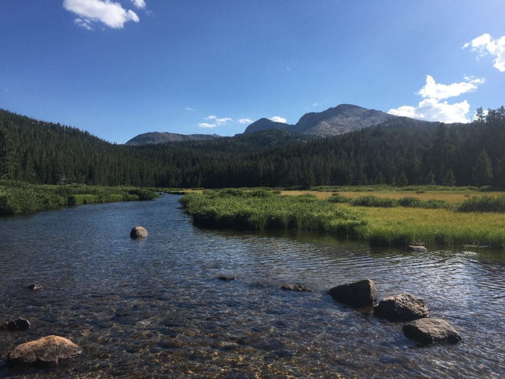 A shallow, clear river winds through a meadow in front of an impressive mountain range.