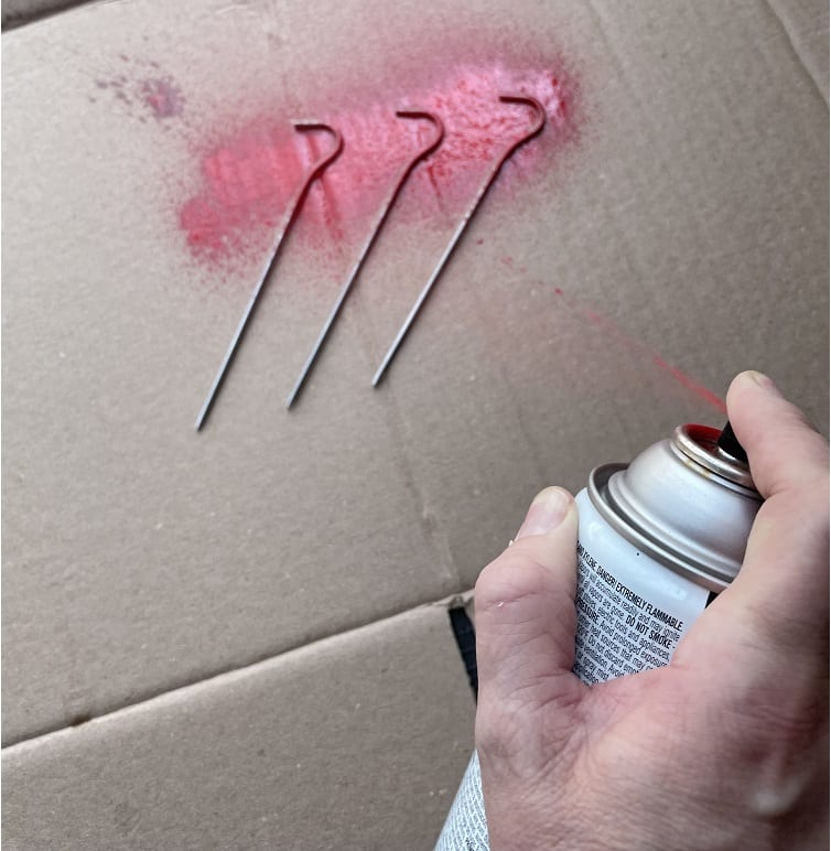 A hand spraying tent stakes over a pice of cardboard with spray paint
