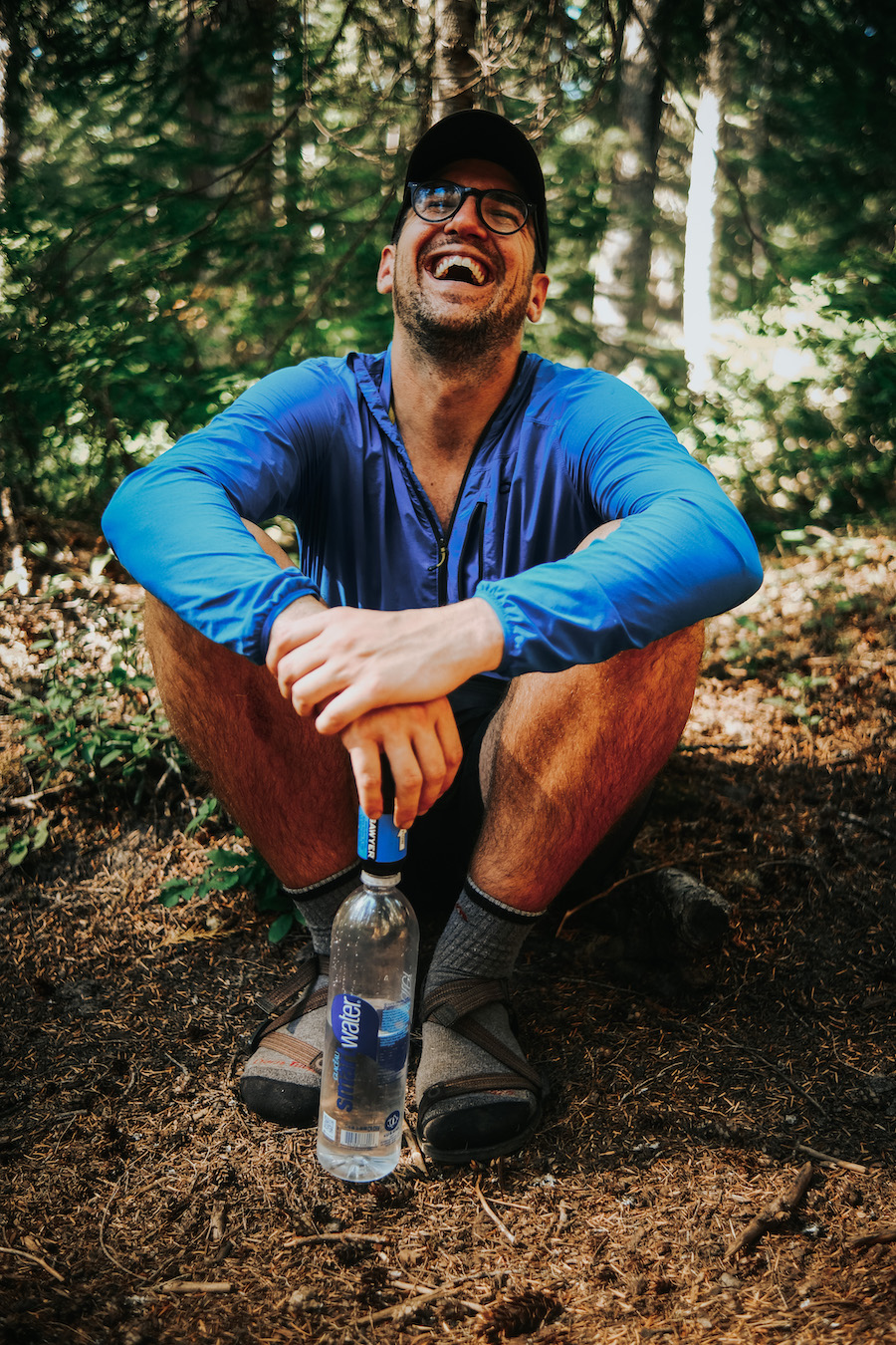 a hiker sitting on the ground laughing while filtering water into his water bottle