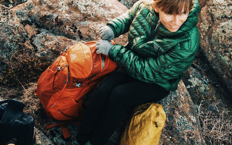 a woman in a green puffy jacket sitting on some rocks opening her backpack