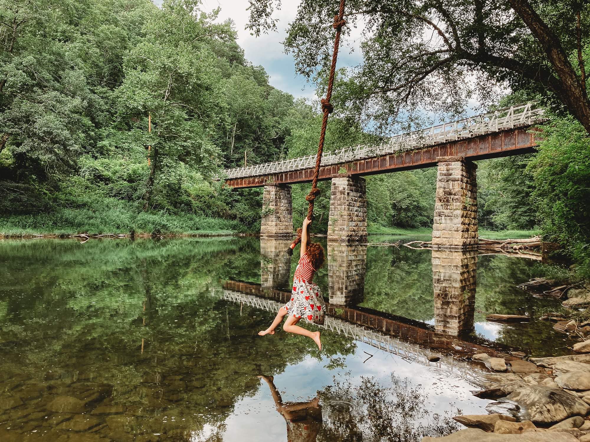 a girl swinging on a rope swing above water on the greenbrier river trail