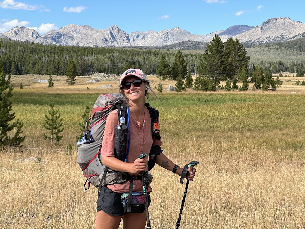 a hiker smiling with mountains and trees in the background