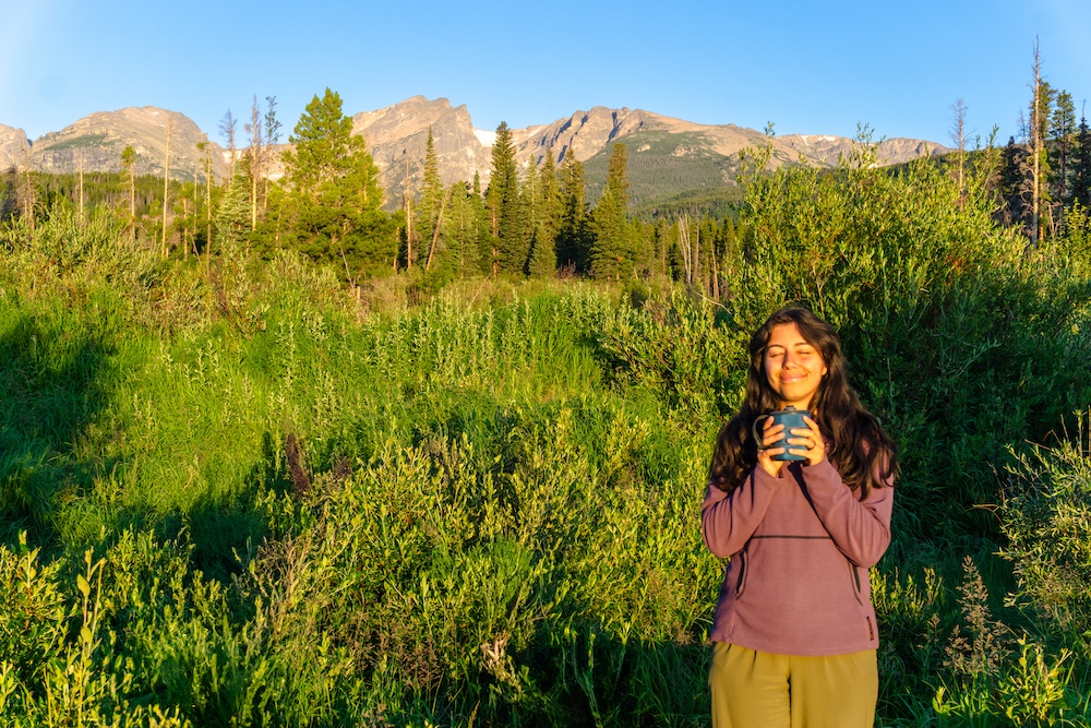a hiker smiling holding a cup with mountains and trees and bushes in the background